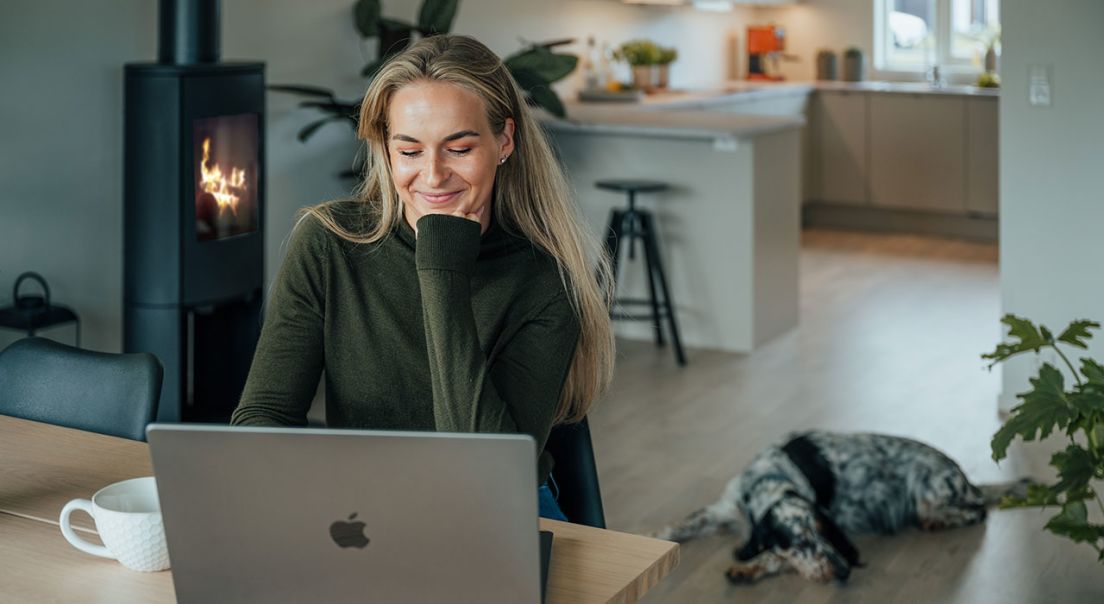 Kvinne sitter på laptop ved spisebord, med hund i bakgrunnen