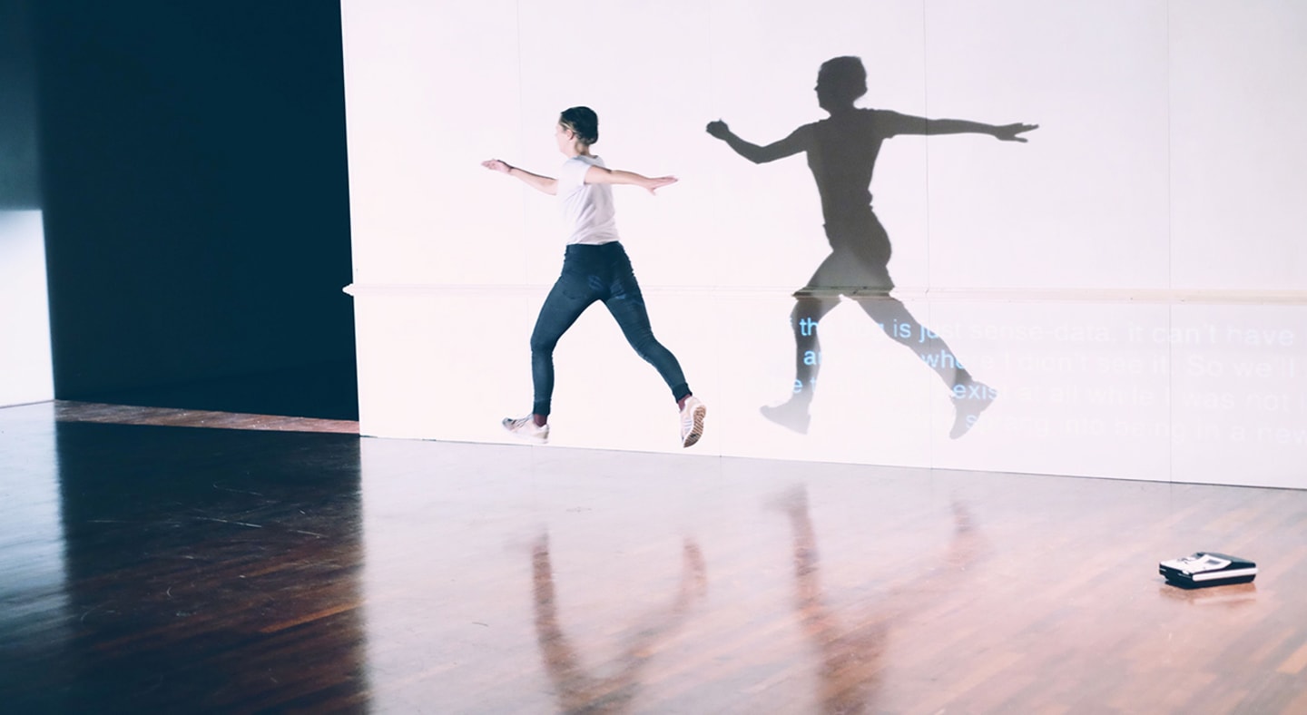 En kvinnelig danser i olabukser og hvit t-skjorte i luftig svev over scenegulvet.