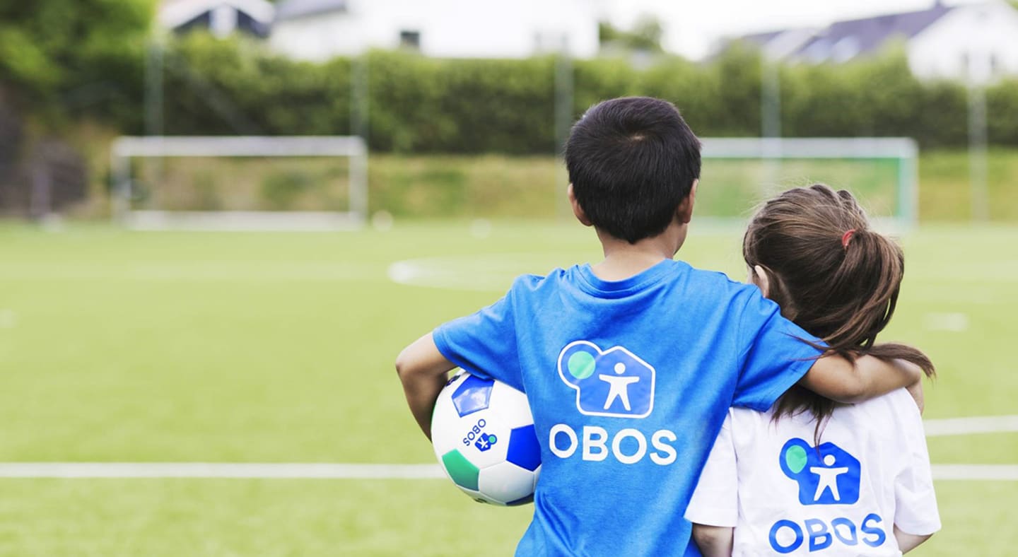 To barn med hver sin OBOS-trøye på seg som holder rundt hverandre mens de ser utover den grønne fotballbanen