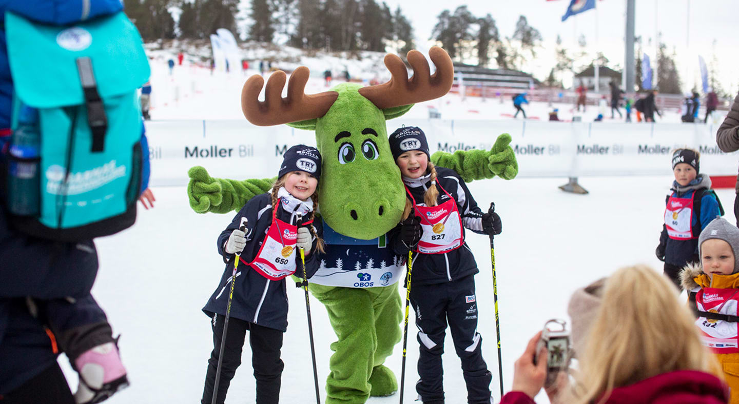 To jenter med ski og turtøy poserer sammen med en av maskotene på Barnas Holmenkolldag.