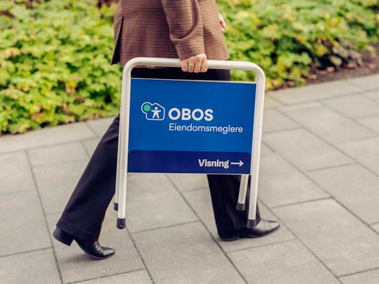 En person går med et visningsskilt for OBOS Eiendomsmeglere på et uteområde.