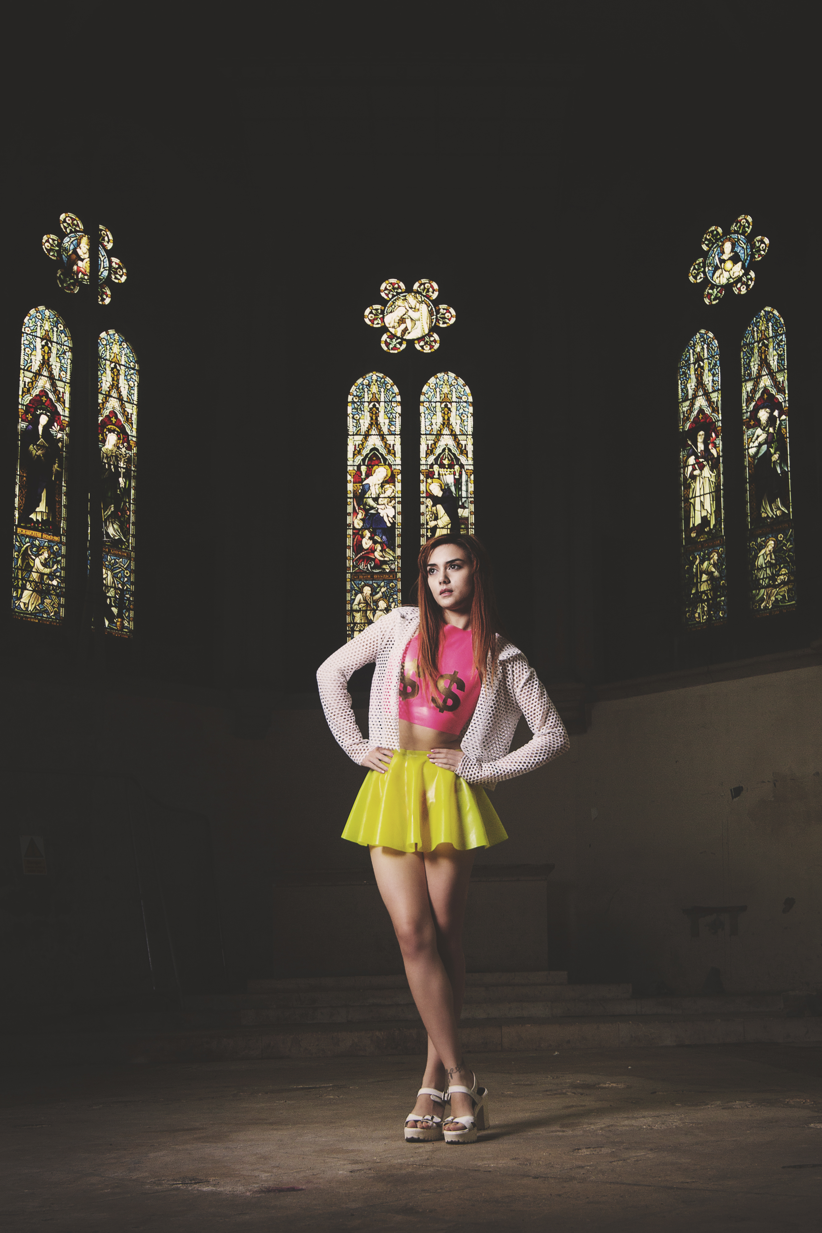 Full-length alternative fashion portrait of a model standing in a dark church interior with towering stained glass windows, captured by Carl Hewlett for Obsidian Noir. She is wearing a neon yellow latex skater skirt, a pink crop top with dollar signs by Shokushu Boutique, and a white mesh jacket.