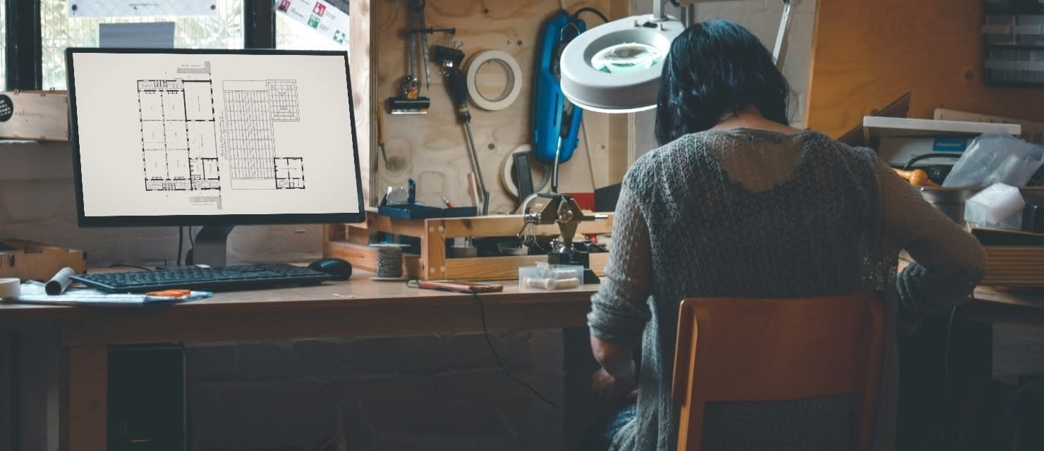 Someone working at a workshop bench, with an architectural floor plan open on the monitor beside them