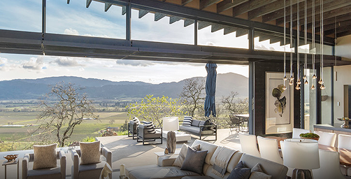 Luxury open-concept living and dining area with exposed wood ceiling beams, floor-to-ceiling glass walls, and folding glass doors opening to a terrace with panoramic vineyard views, featuring modern furniture and pendant lighting.