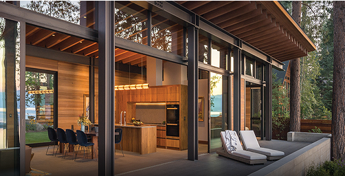 Modern lakefront home with exposed wood ceiling beams, floor-to-ceiling glass walls, open-concept kitchen with wood cabinetry, and outdoor patio featuring lounge chairs overlooking serene water views.