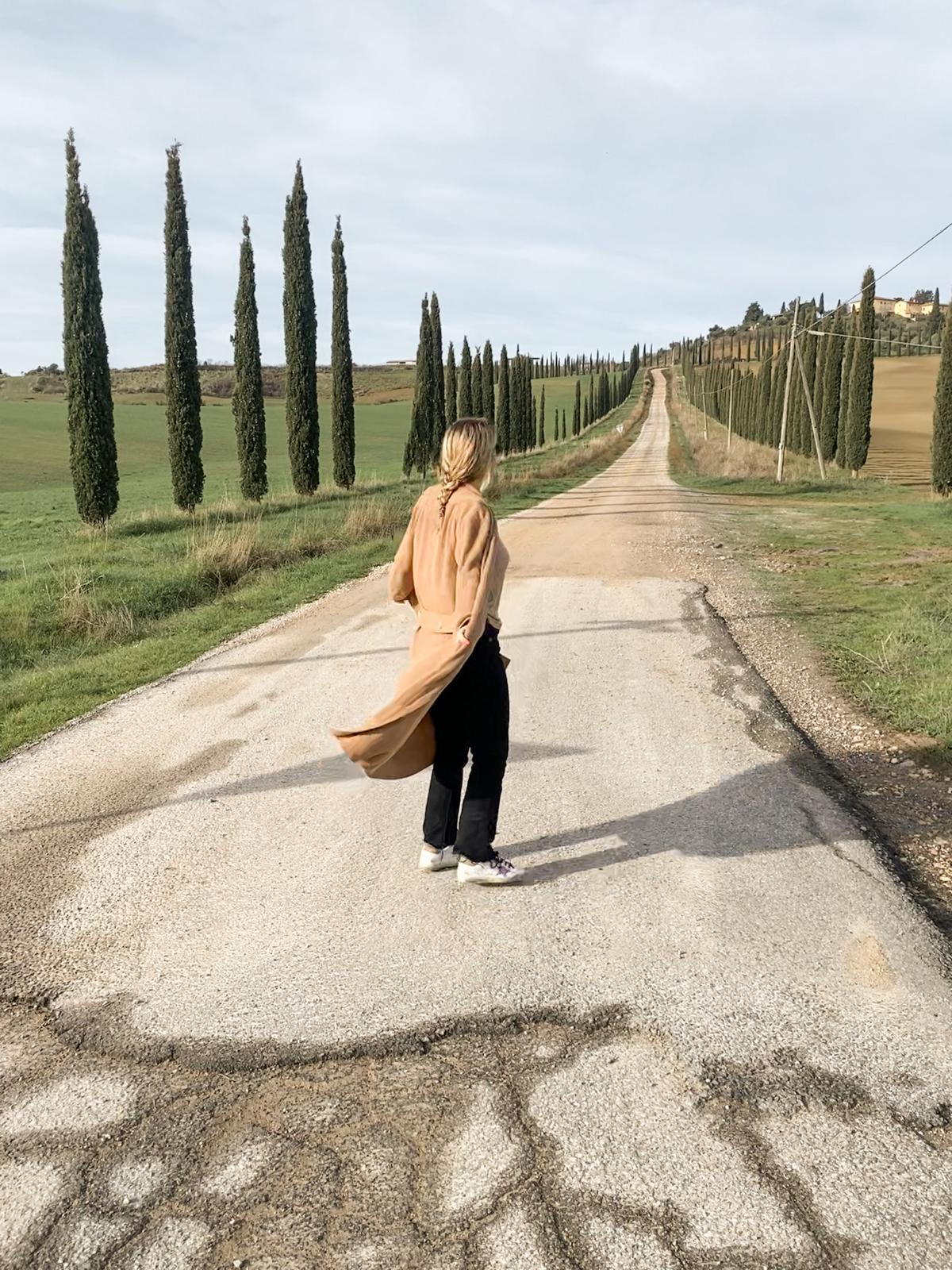 A woman in a camel coat walking away down a cypress-lined gravel road