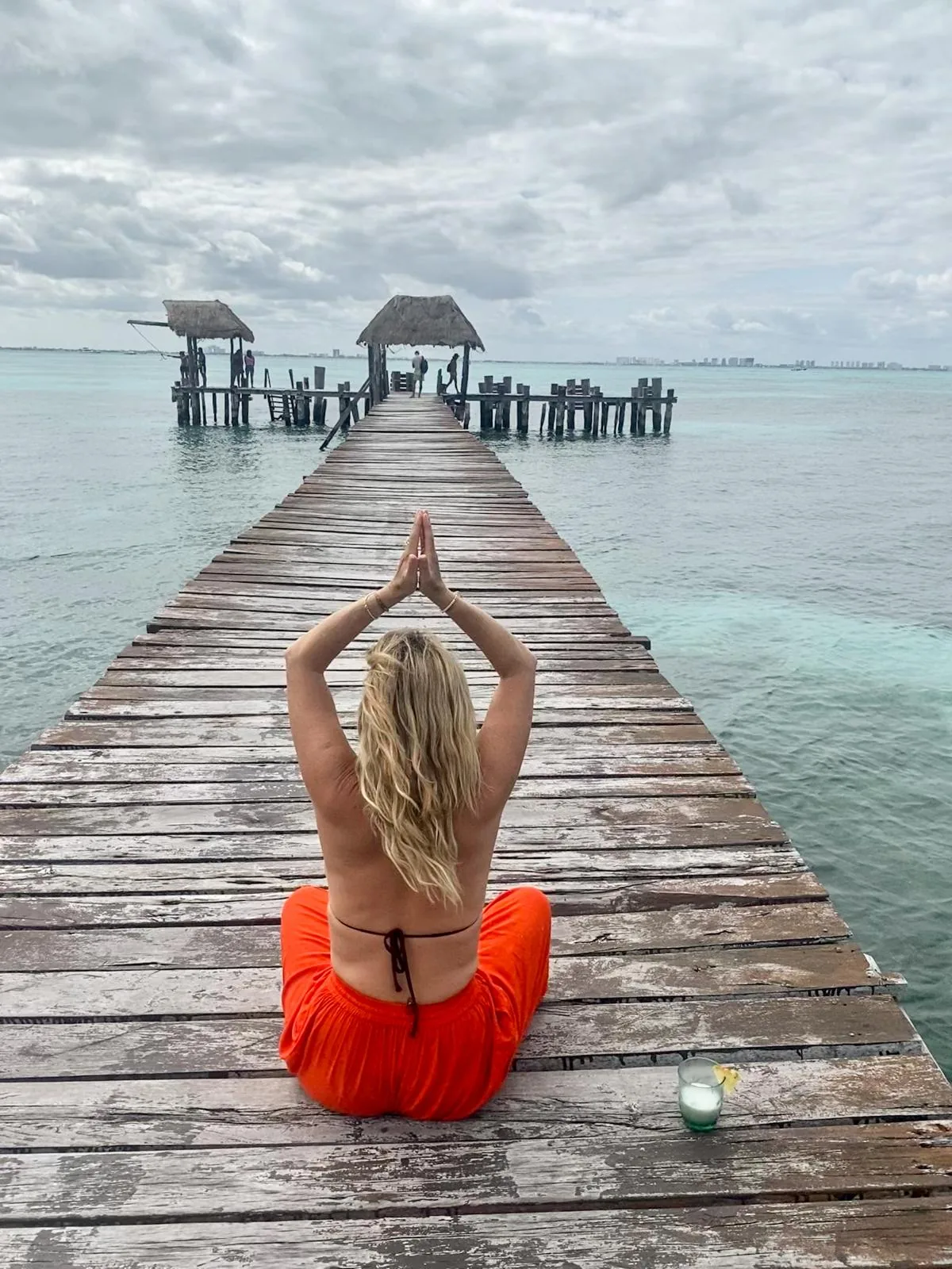 A woman in an orange dress seated on a wooden jetty over turquoise water, hands raised together