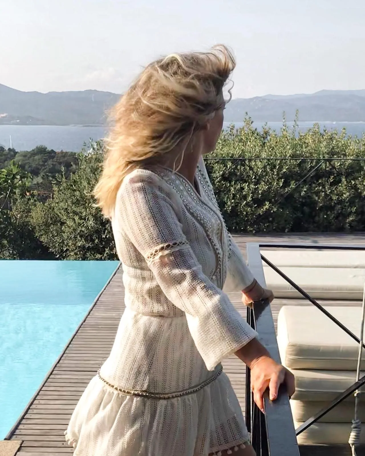 A woman in a white dress at the edge of an infinity pool, hair lifted by the wind