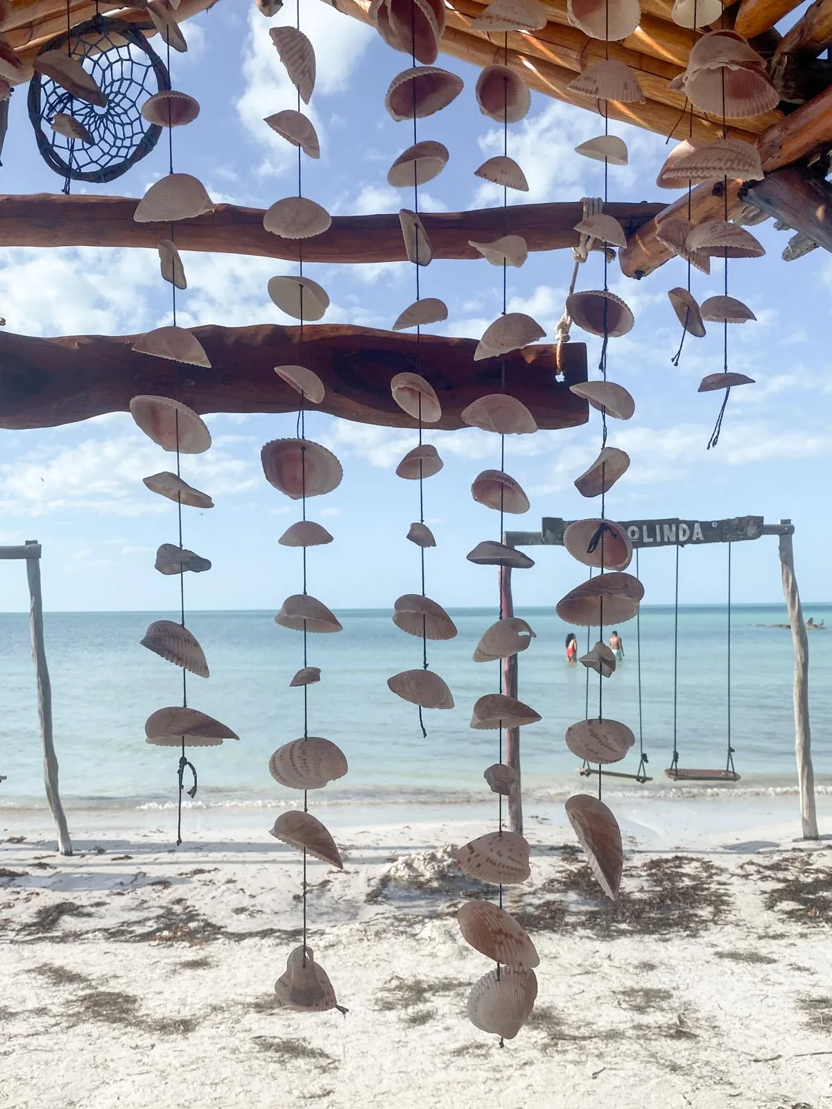 Hanging shell mobiles under a driftwood shelter above a white beach