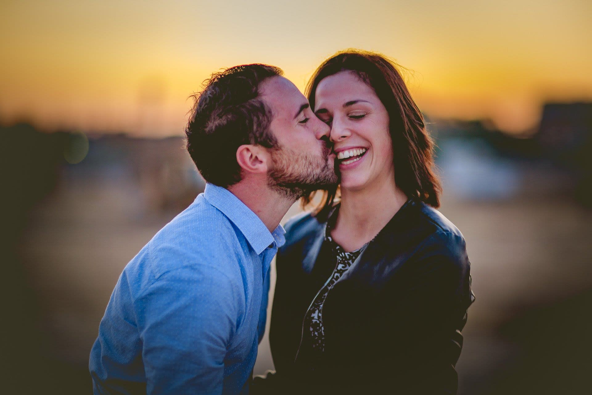 He kisses her on the cheek while she laughs, the golden sunset sky behind them