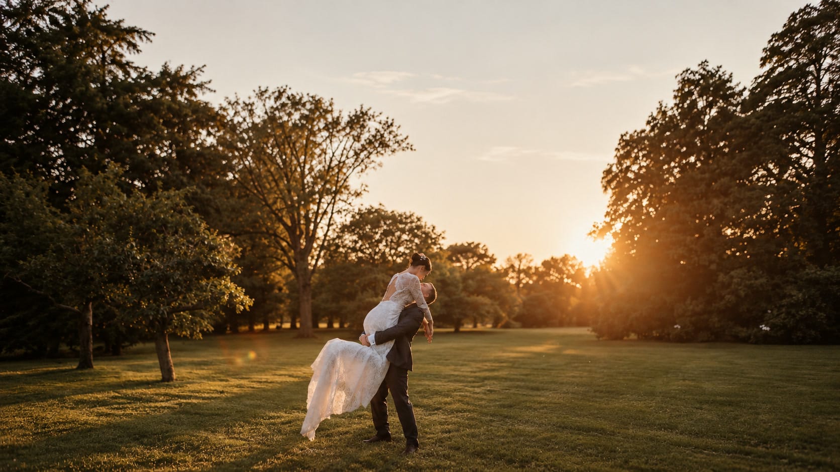 The groom lifting the bride on a park lawn, backlit among the trees at sunset