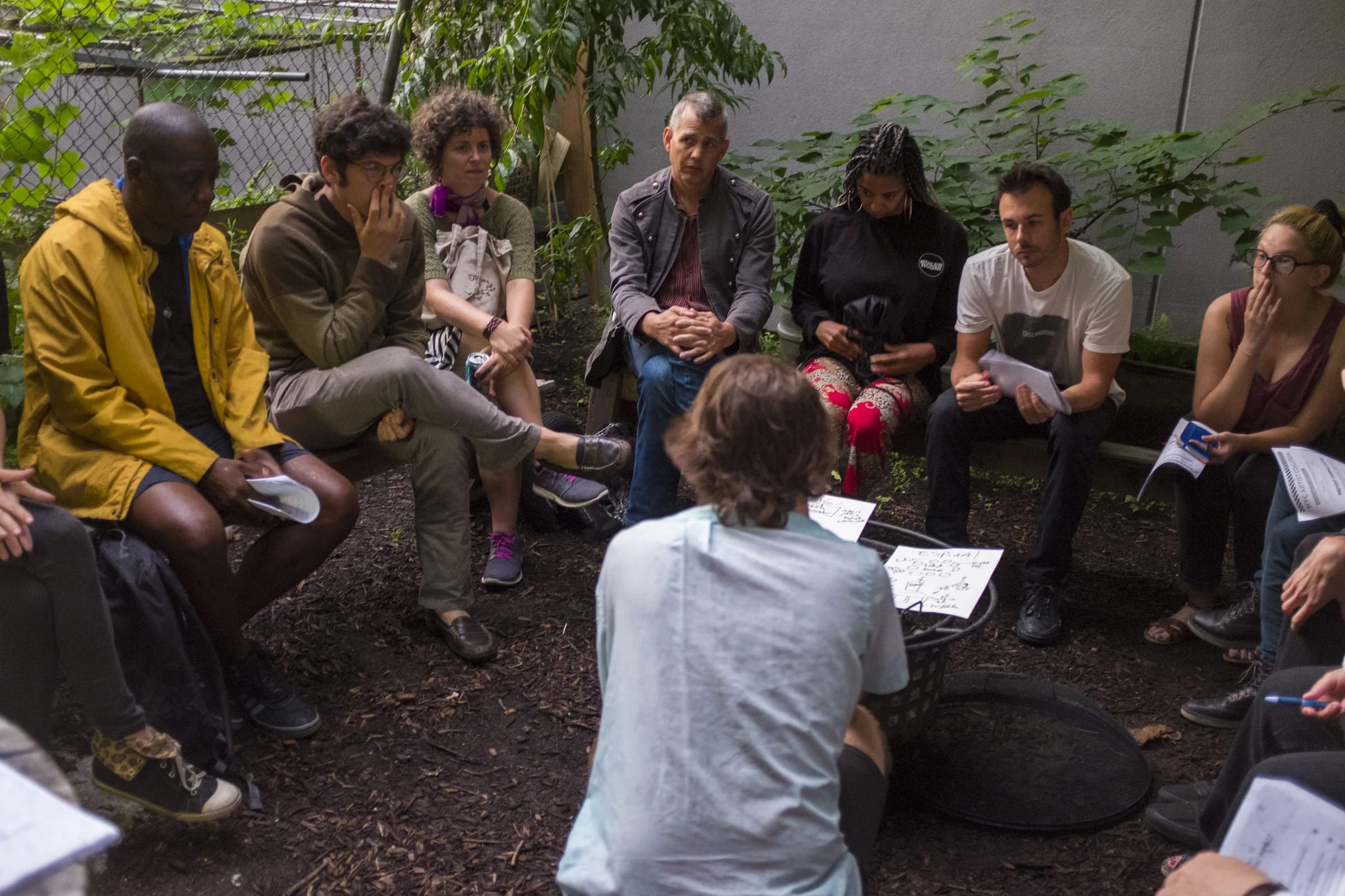 Jamie Burkart, seen from behind, crouches in a garden circle while participants look toward shared handwritten notes on a low grill.