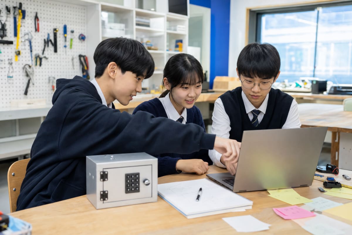 한국 학교 메이커실에서 노트북과 프로젝트 노트를 보며 AI 소프트웨어 공모전을 준비하는 청소년 세 명