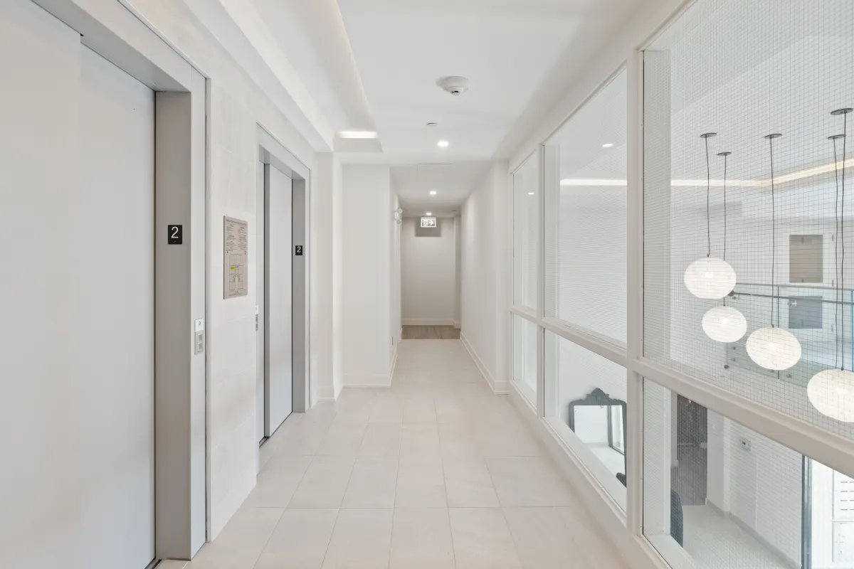 Image of a white hallway, with an elevator on the left side and on the right a window that looks into a room with a mirror leaning on a wall and a desk with some lights hanging from the roof. The goal of this image is to think about hotels and other such appartment buildings.