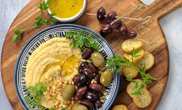 EN tallerken med hummus, oliven og pinjekjerner dandert på en trefjøl med olivenolje og crustini.