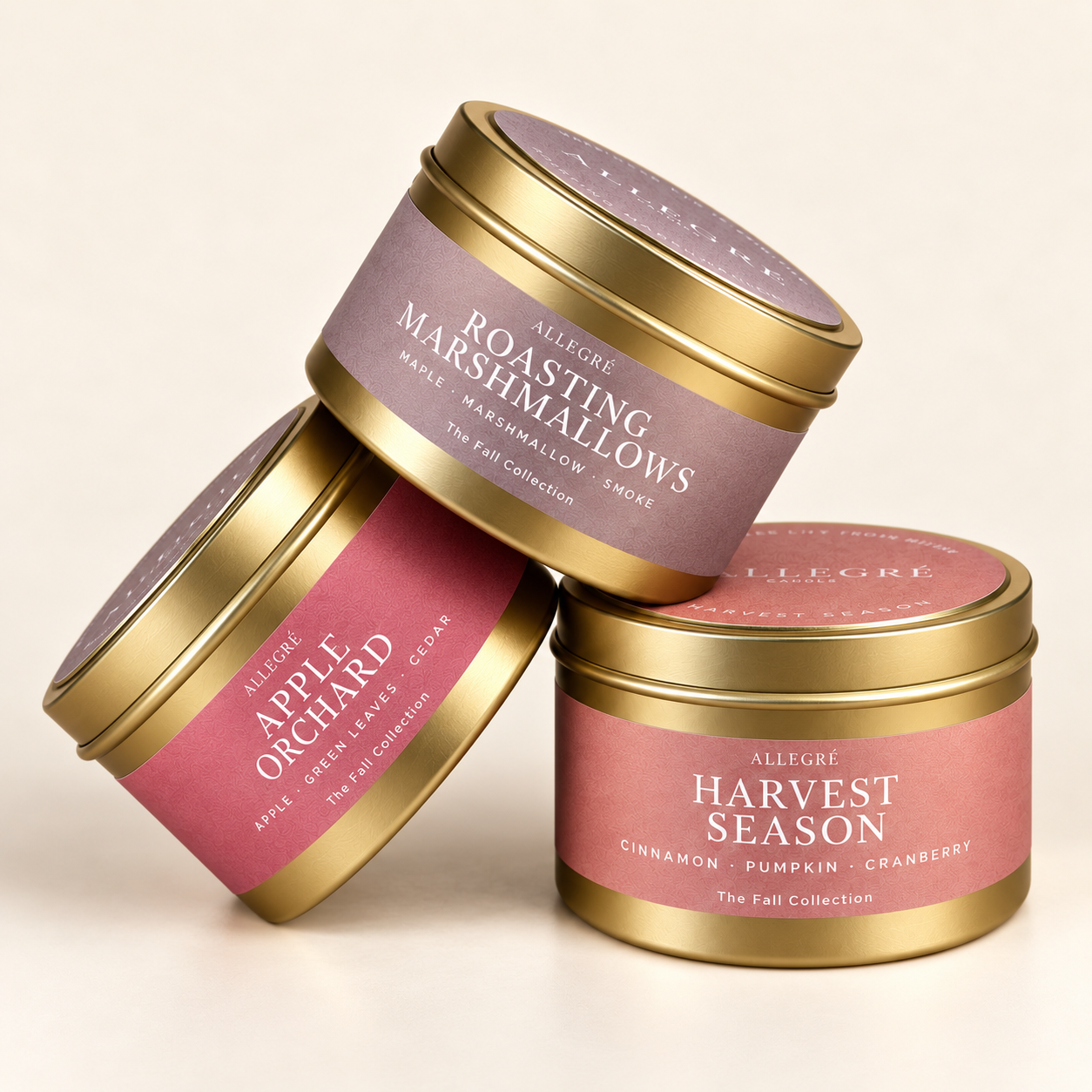 Three ALLEGRE Fall Collection coconut-soy candles in gold tins with pink and mauve labels, featuring Roasting Marshmallows, Apple Orchard, and Harvest Season.