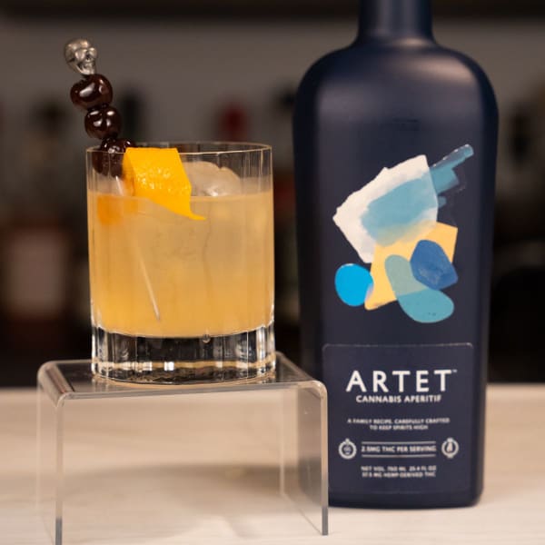 Cannabis smoked old fashioned with a skull cherry pick and orange twist beside a bottle of Artet cannabis aperitif