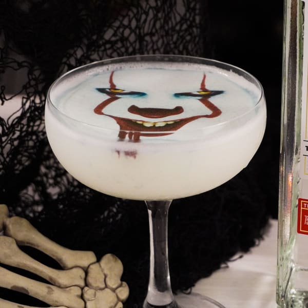 White Lady gin cocktail in a coupe with an edible image of Pennywise the Clown floating on the egg white foam, beside a skeleton hand and black netting