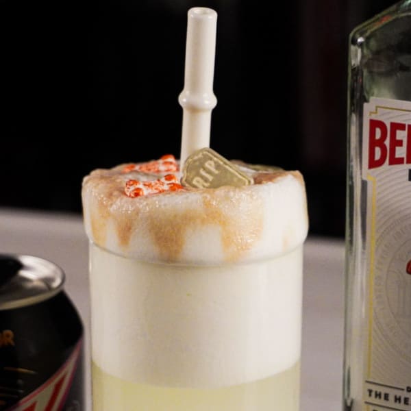 Halloween Ramos gin fizz in a Collins glass with a tall foam column above the rim, a bone straw standing upright in it, a tombstone decoration, and a Beefeater bottle behind