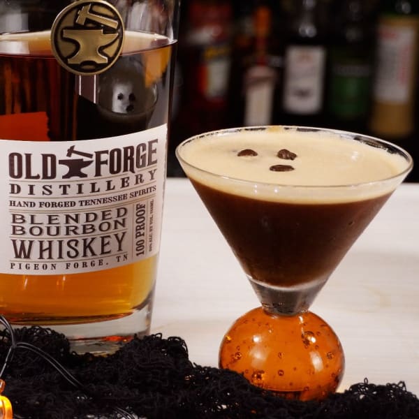 Pumpkin spice espresso martini with a three coffee bean garnish in a bubble-stem martini glass, beside a bottle of Old Forge Distillery blended bourbon from Pigeon Forge, Tennessee