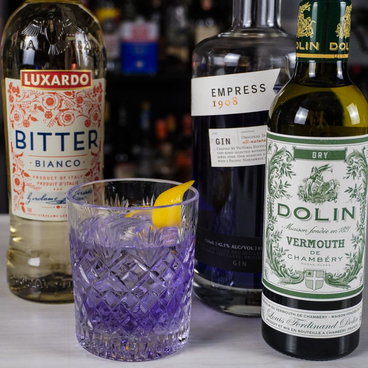 Purple Negroni in a cut crystal rocks glass with a lemon peel, in front of Luxardo Bitter Bianco, Empress 1908 gin, and Dolin dry vermouth