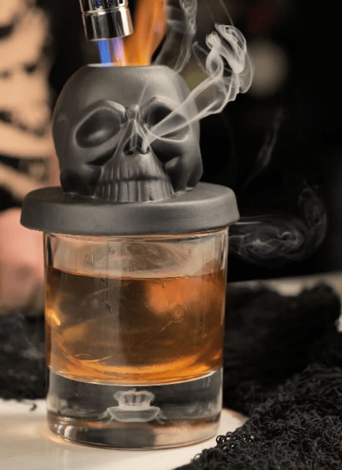 Smoke pouring from the skull cocktail smoker while torching wood chips over a smoked old fashioned