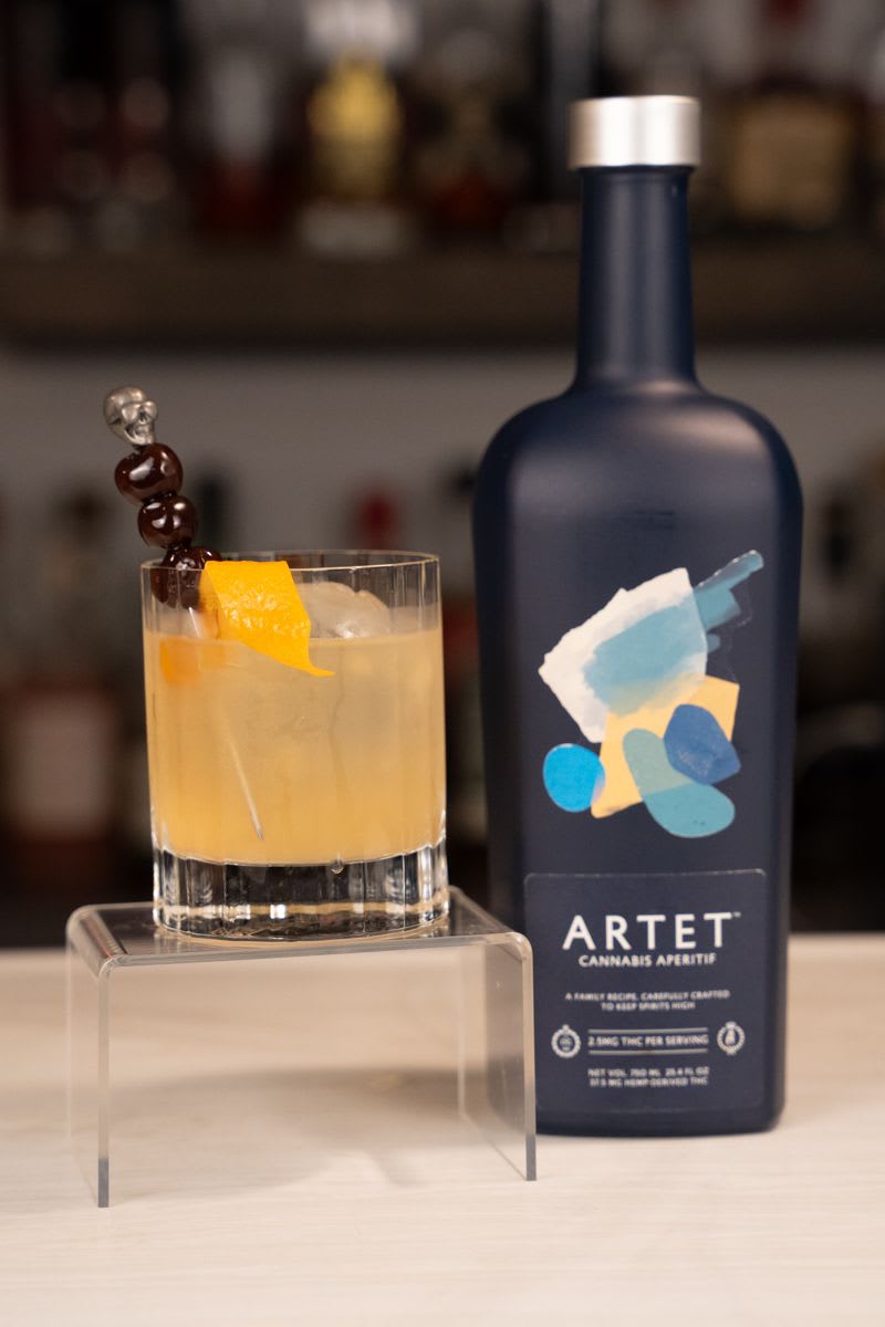 Cannabis smoked old fashioned with skull cherry pick and orange twist next to a bottle of Artet cannabis aperitif