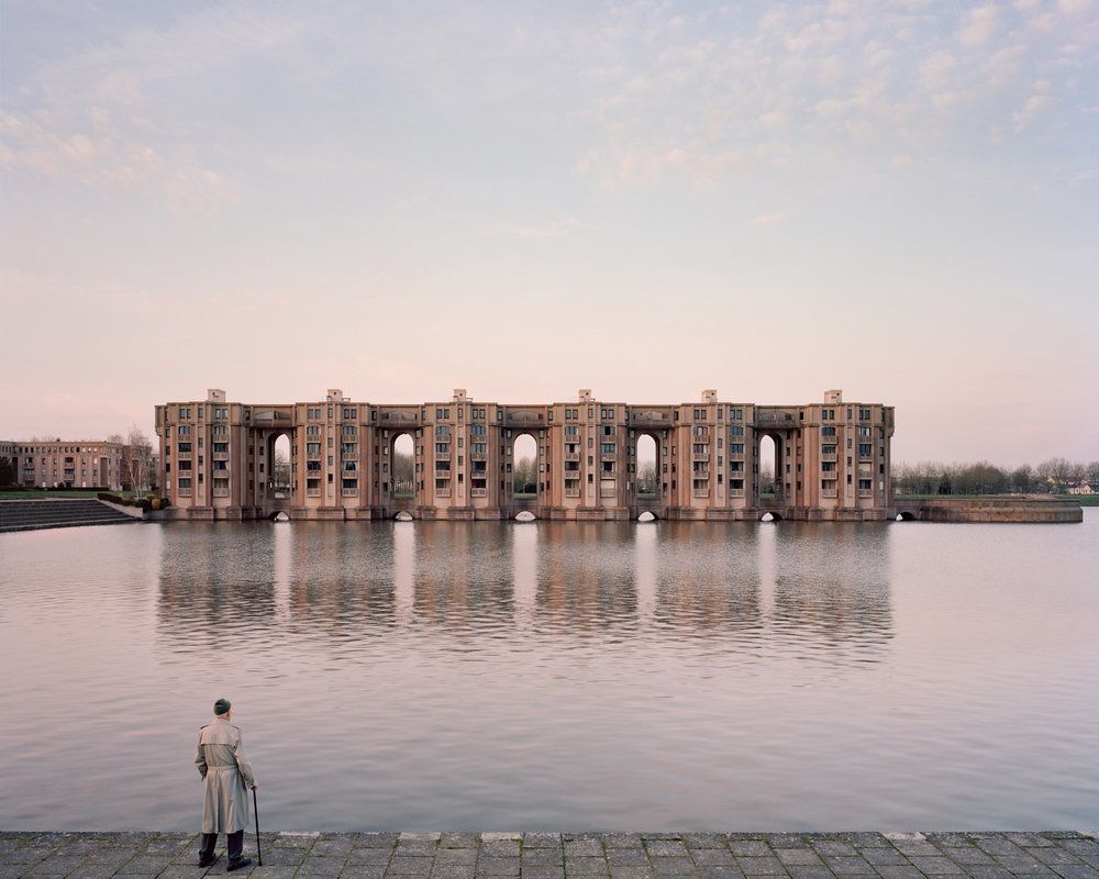 Photograph of a man with a cane, standing in front of a body of water, looking at a housing estate in the distance