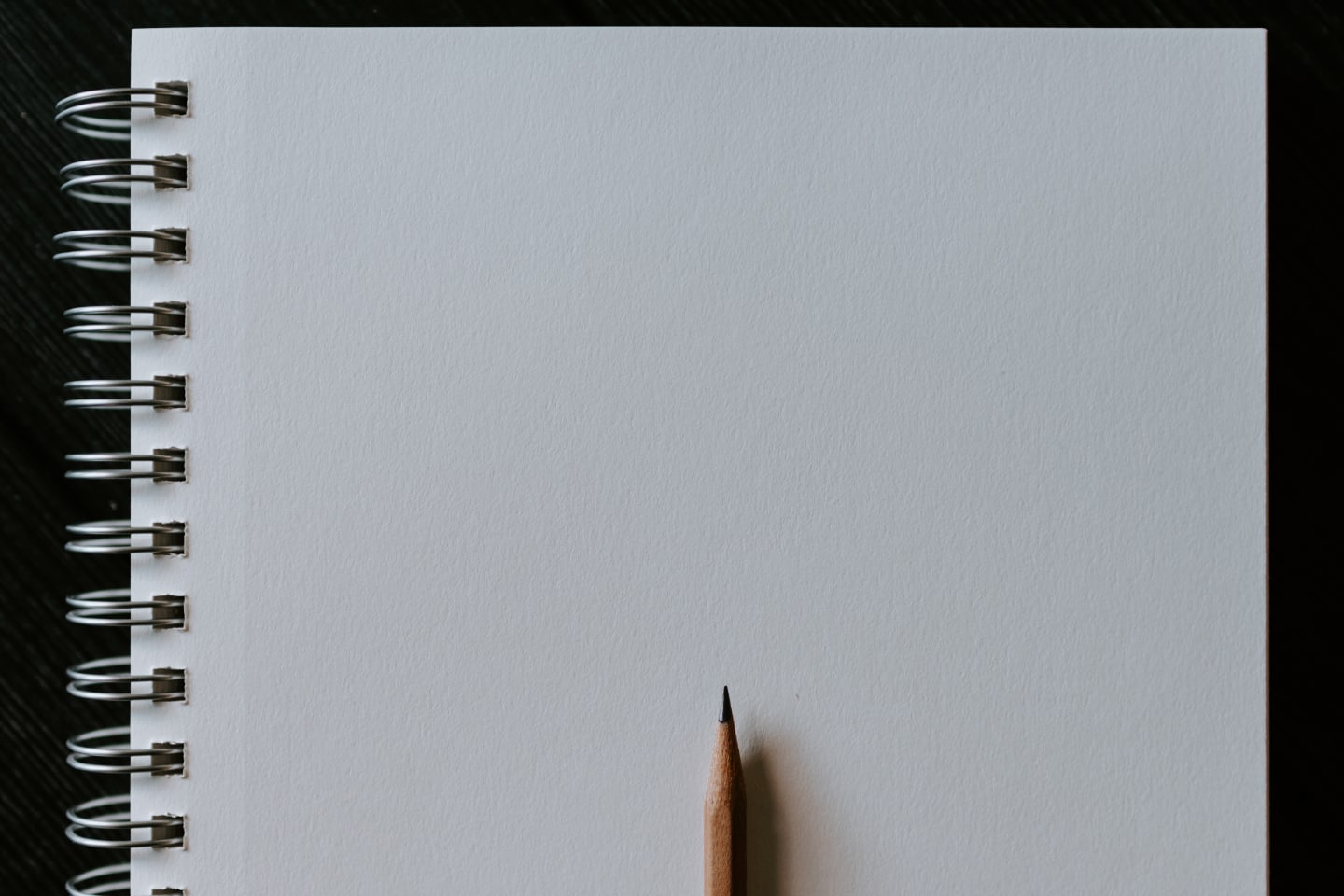 A blank, white page in a spiral-bound notebook with a sharpened pencil laying on it.