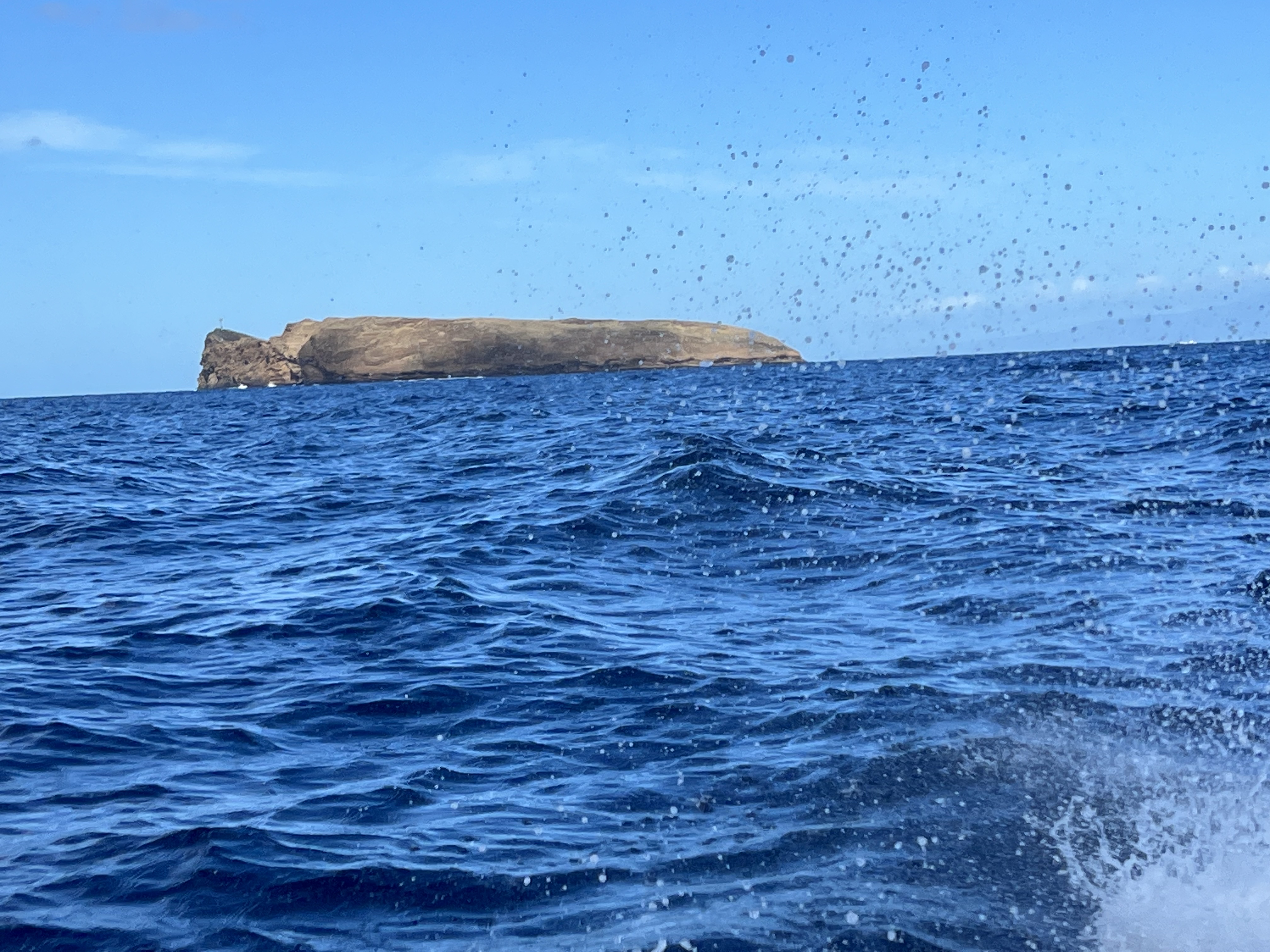 Molokini Crater seen from the water on the boat ride out — the crescent rim rising from a deep blue channel, sea spray in the air