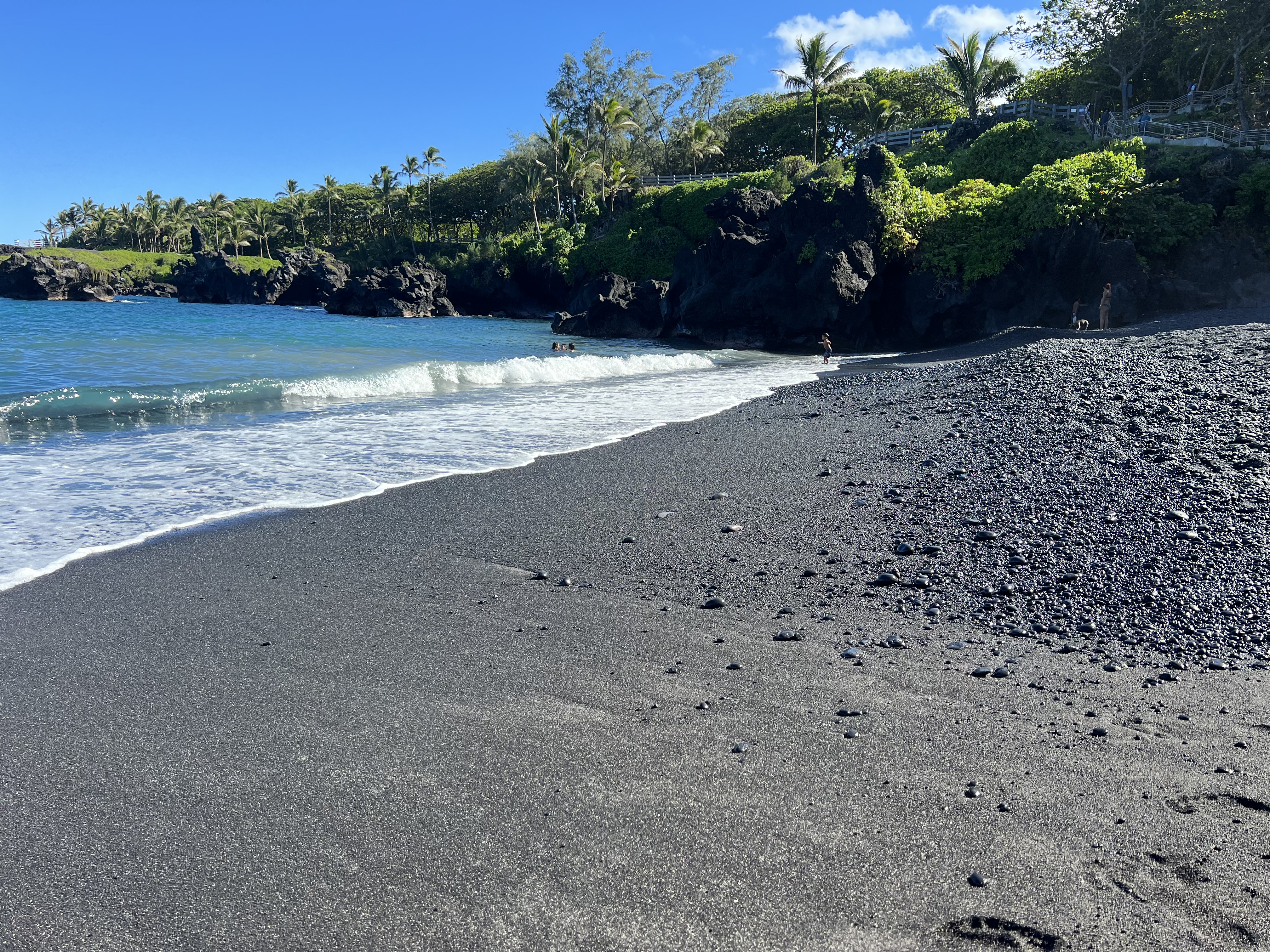 Wai'anapanapa's black sand beach at shoreline level — jet-black pebbled sand meeting a turquoise wave, palms and lava cliffs behind