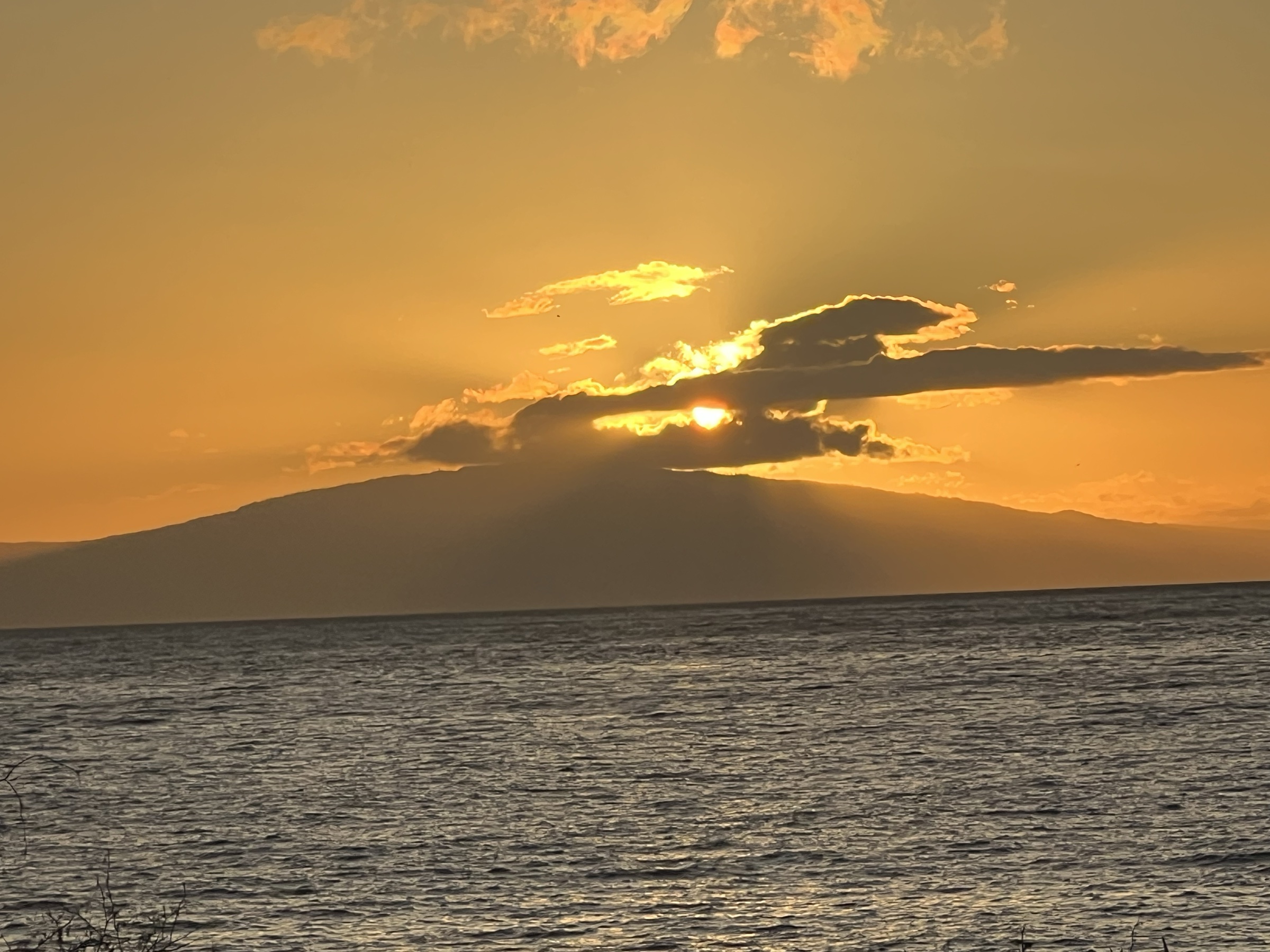 Sun setting behind Lāna'i island with golden rays breaking through clouds