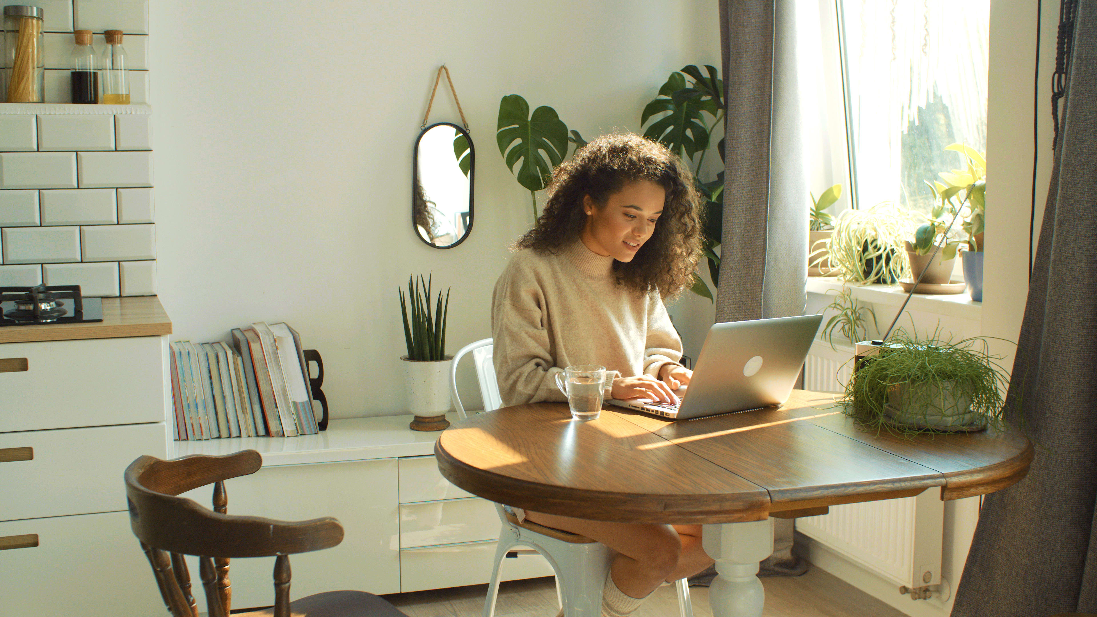 Beautiful young brunette woman sitting in cozy kitchen by a table and working on her laptop computer.