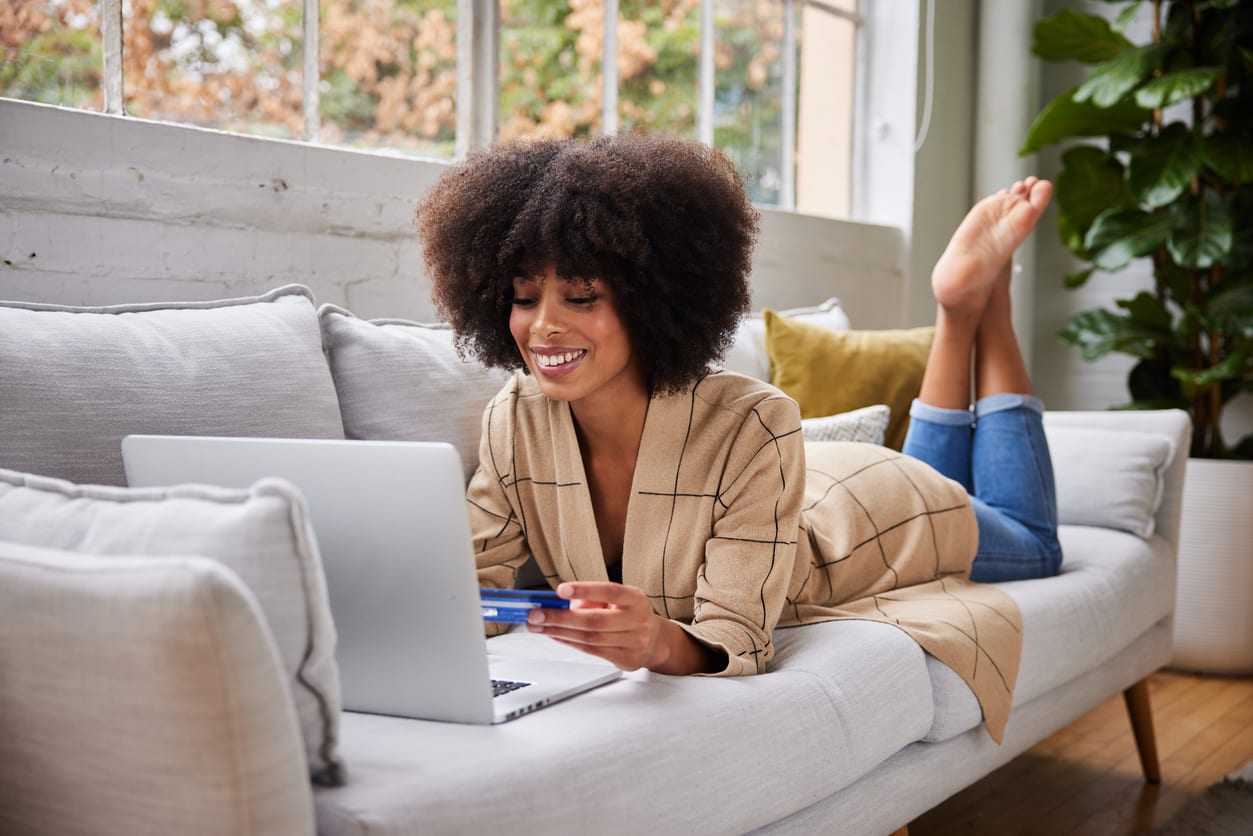 Smiling young woman lying on her living room sofa shopping online with a credit card and laptop