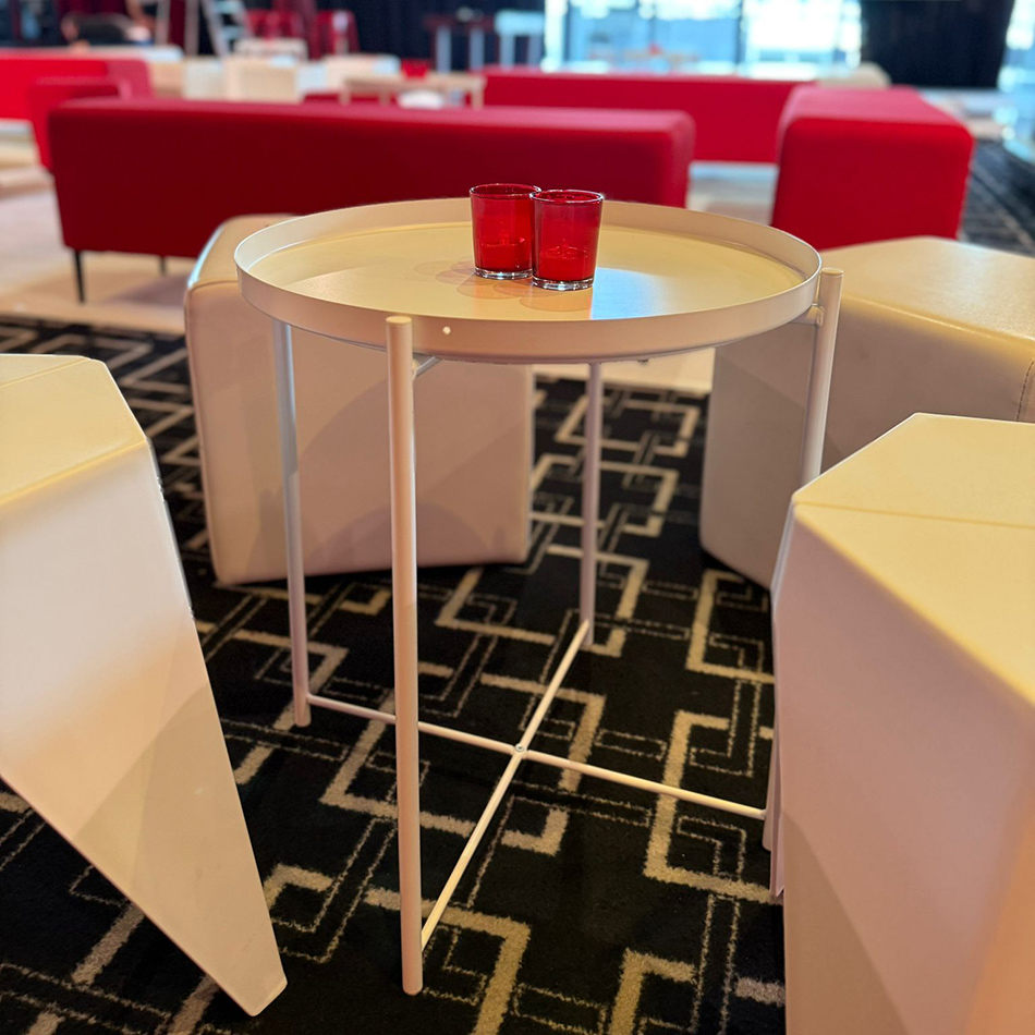 Close-up of a white tray side table with two red votive candles, surrounded by white Puzzle Stools and cube ottomans on a patterned black carpet in a modern event space.
