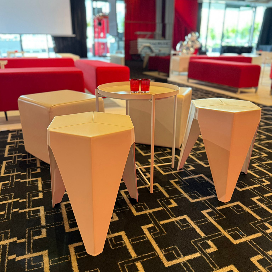 White tray side table with red glass candles centred among white Puzzle Stools and ottomans on a geometric carpet, with red lounge seating in the background at a stylish indoor venue.