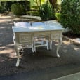 Cream vintage table with decorative handles and curved legs, positioned with two white chairs on a paved garden path surrounded by greenery.