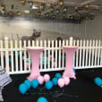 A room decorated with baby pink balloons and a white picket fence.