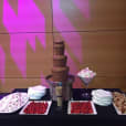 A chocolate fountain on a table with 1kg of strawberries and desserts on it.