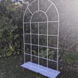 A white arbour, resembling a French window arch frame, situated in the middle of a garden.