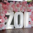 A pink and white balloon arch with the name Zoe illuminated by glow letters.