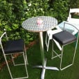 A stainless steel cocktail bar table and two stools in a yard.