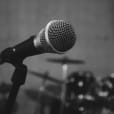 A black and white photo of a corded microphone in front of a drum set.