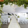 A wedding ceremony set up in a tent with white flowers and greenery, featuring elegant White Padded Folding Chairs (Gladiator chair) for seating.