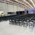 A large warehouse with rows of black padded folding chairs and a large screen.