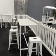 A room adorned with a white picket fence and white stools surrounding a White Rectangular Tapas Table Hire with a white top.