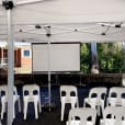 A white tent with white chairs and a projector screen hire.