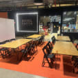 Event setup featuring multiple black hairpin banquet tables with natural timber tops, paired with black folding chairs on an orange floor covering, arranged in a retail venue space for a workshop or gathering.