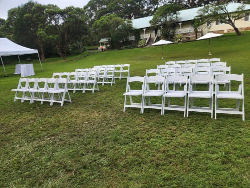 Scores of white folding plastic chairs in an outdoor park next to a marquee