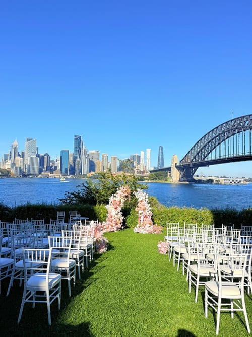 Lots of white Tiffany chairs set up for a wedding in a park in front of the Harbour Bridge